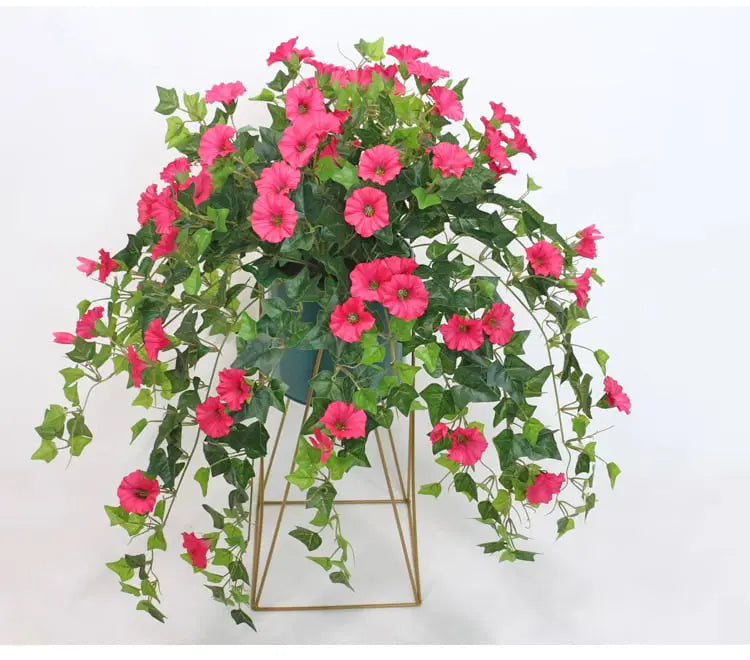 Roze kunstbloemen met groen blad die over de rand van een donkere pot op een gouden metalen standaard hangen. 3