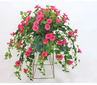 Roze kunstbloemen met groen blad die over de rand van een donkere pot op een gouden metalen standaard hangen. 3