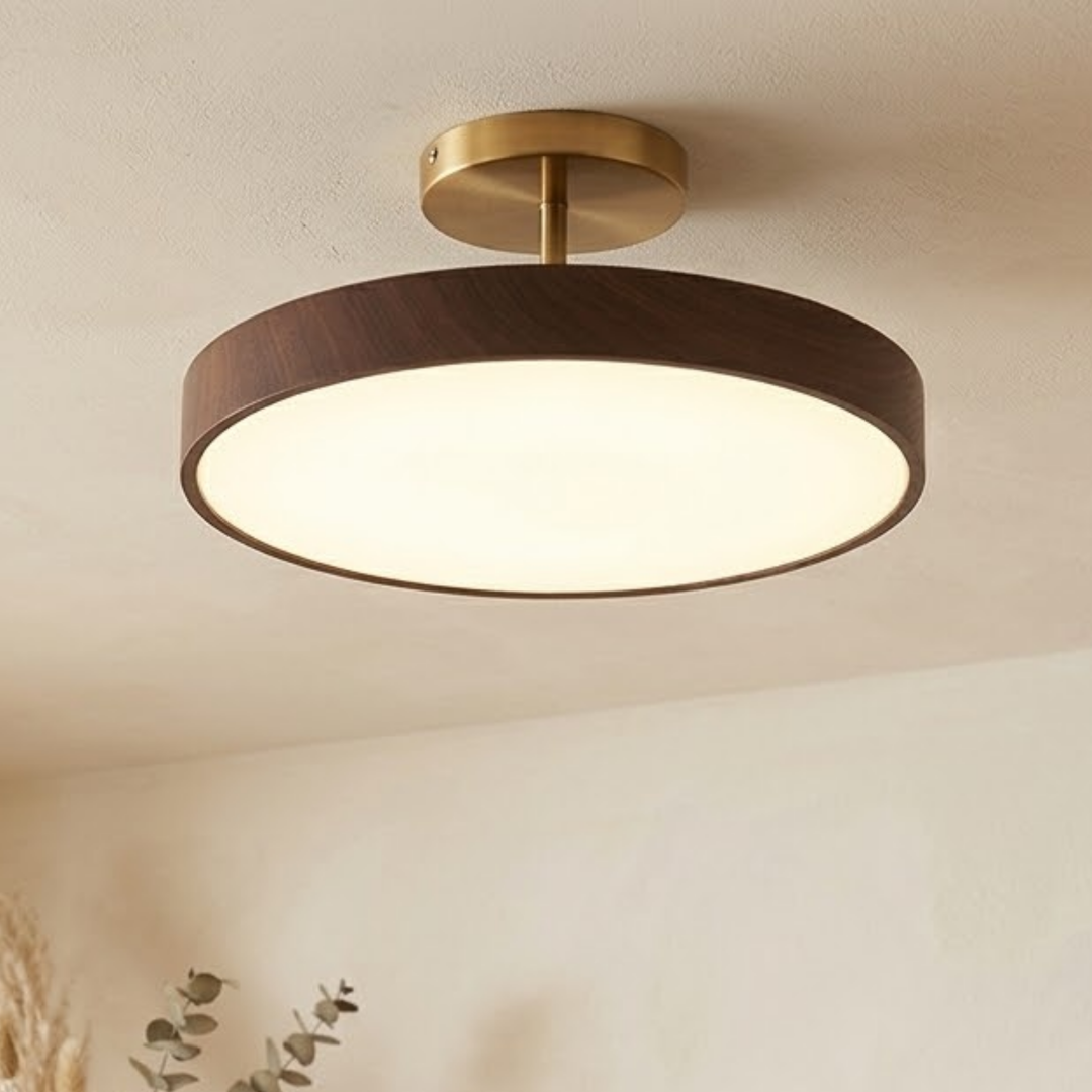 Ronde plafondlamp met donkere houtlook rand en goudkleurige steun, brandend aan een licht plafond met eucalyptustakjes eronder. 1