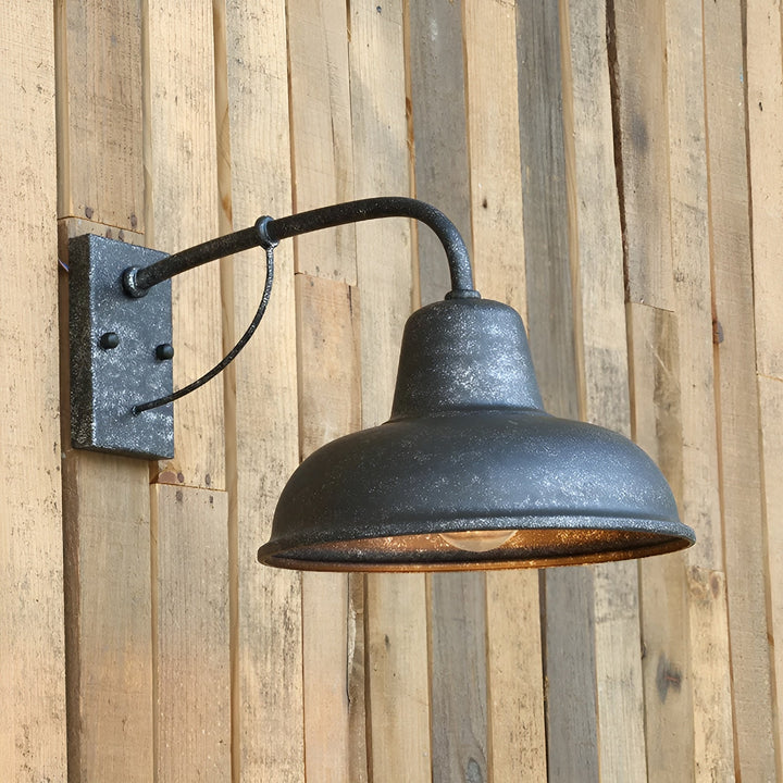 Wandlamp met verweerde, gespikkelde donkergrijze afwerking bevestigd tegen een houten plankenwand, met brandende gloeilamp onder de kap. 2