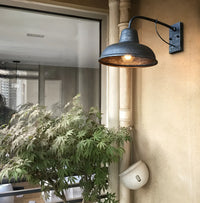 Wandlamp met gespikkelde donkere afwerking aan een beige buitenmuur naast een regenpijp en een plant, met brandende lamp. 3