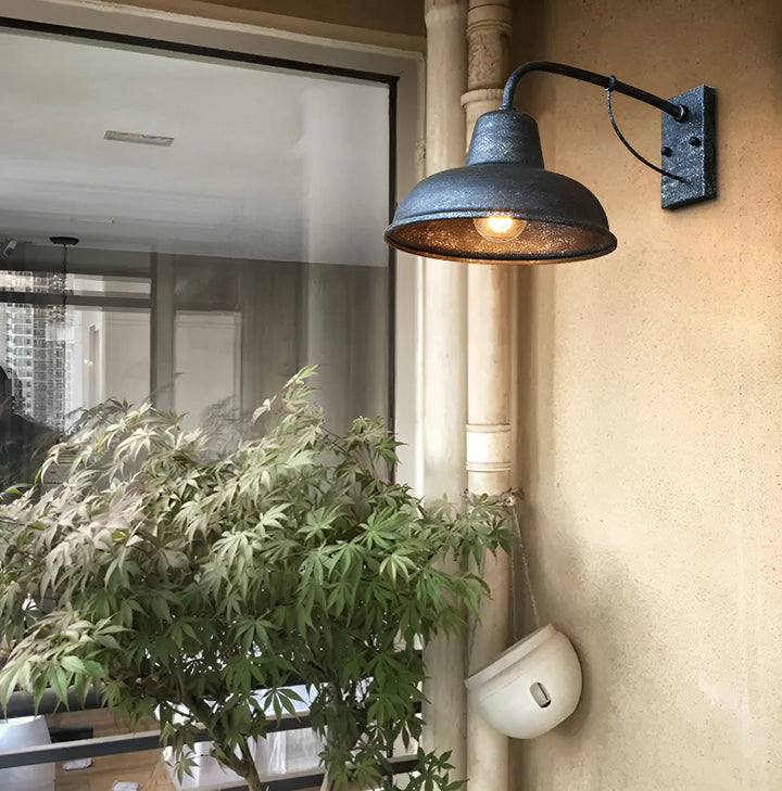 Wandlamp met gespikkelde donkere afwerking aan een beige buitenmuur naast een regenpijp en een plant, met brandende lamp. 3