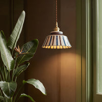 Gestreepte keramische hanglamp met warm licht naast een grote groene plant met bloem, voor een bruine muur. 4