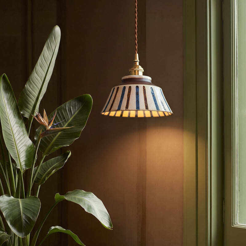 Gestreepte keramische hanglamp met warm licht naast een grote groene plant met bloem, voor een bruine muur. 4