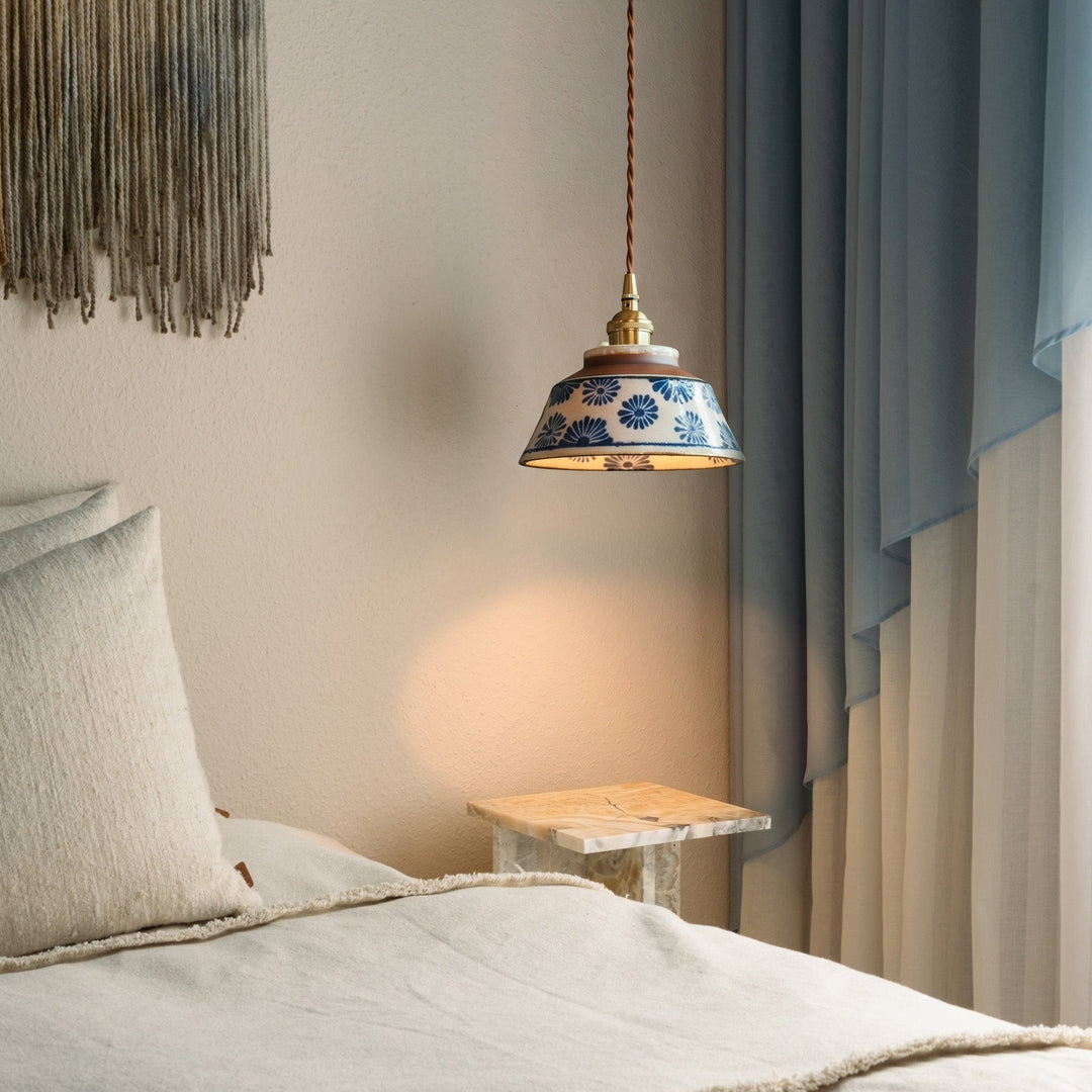 Brandende hanglamp met blauw bloemendecor boven een marmeren bijzettafel naast een bed met linnen kussens en blauwe gordijnen. 2