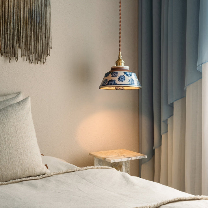 Brandende hanglamp met blauw bloemendecor boven een marmeren bijzettafel naast een bed met linnen kussens en blauwe gordijnen. 2