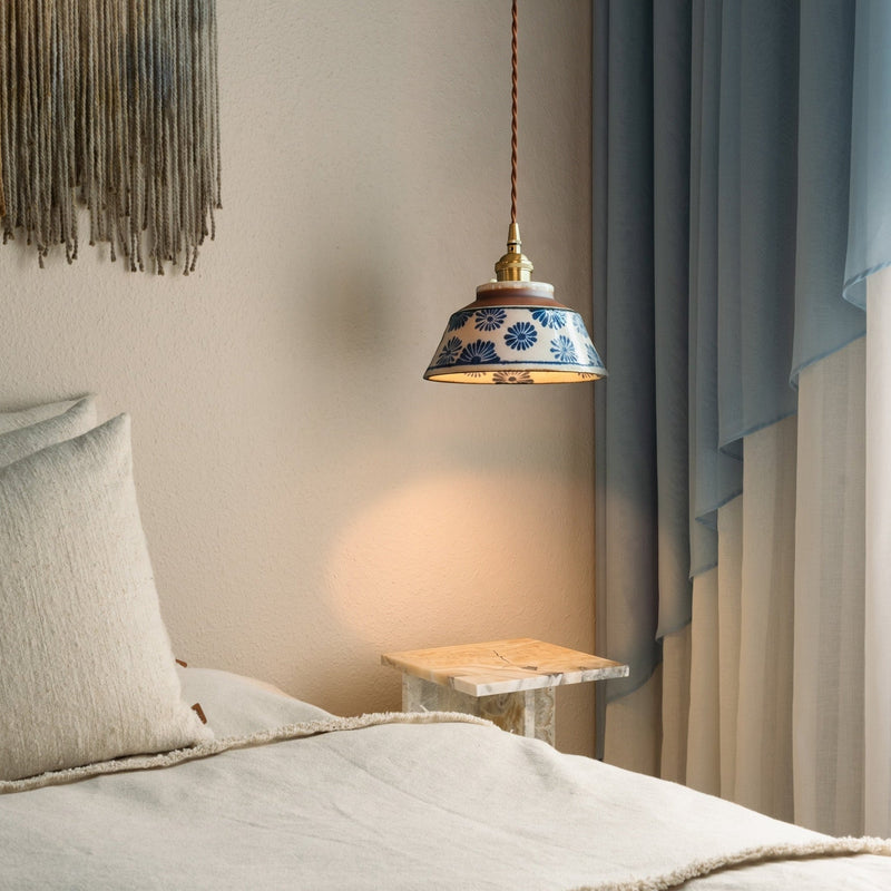 Brandende hanglamp met blauw bloemendecor boven een marmeren bijzettafel naast een bed met linnen kussens en blauwe gordijnen. 2