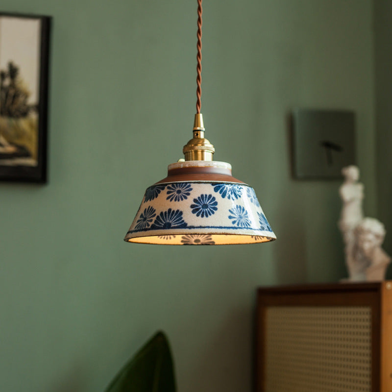 Hanglamp met blauw bloemenpatroon op de keramische kap en messing fitting, brandend voor een groene muur met beeldje. 1