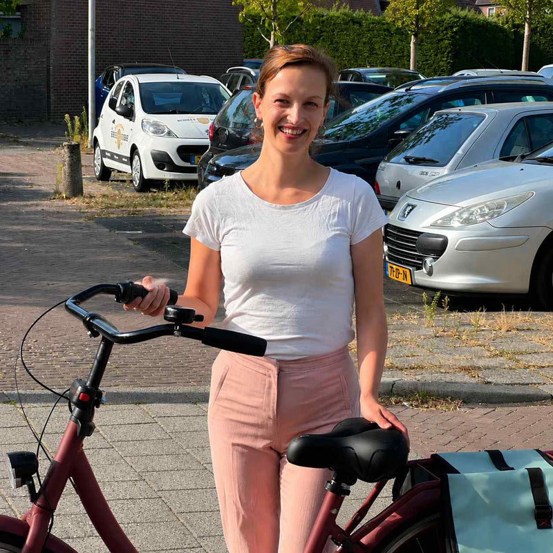 Lachende vrouw staat naast haar fiets met een hand op het zwarte zadel, een fietstas achter het zadel. 7