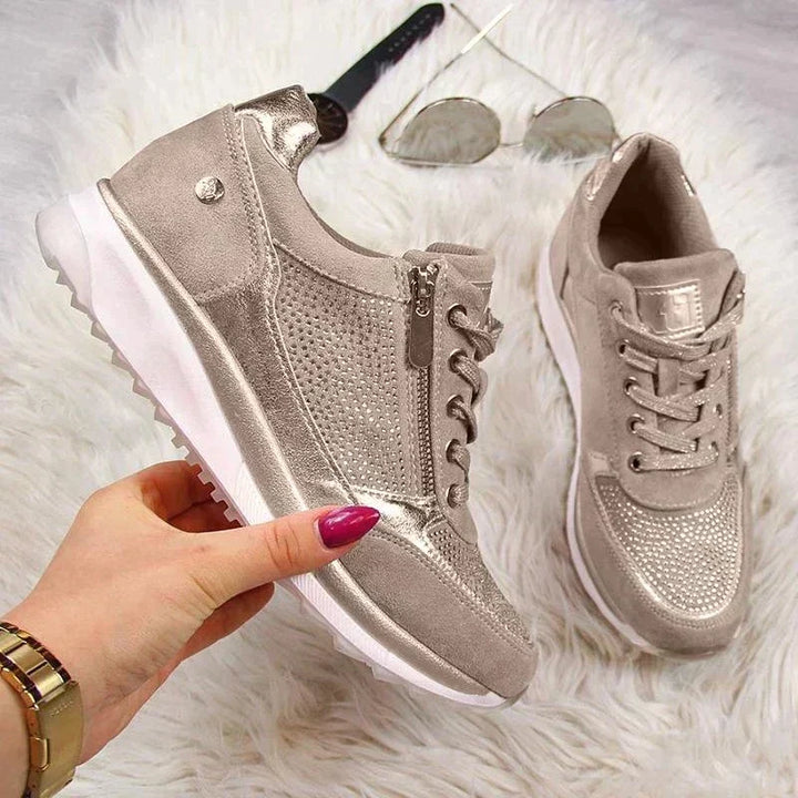 Hand houdt een taupe sneaker met zijrits vast, met de tweede schoen en een zonnebril op wit bont op de achtergrond. 7
