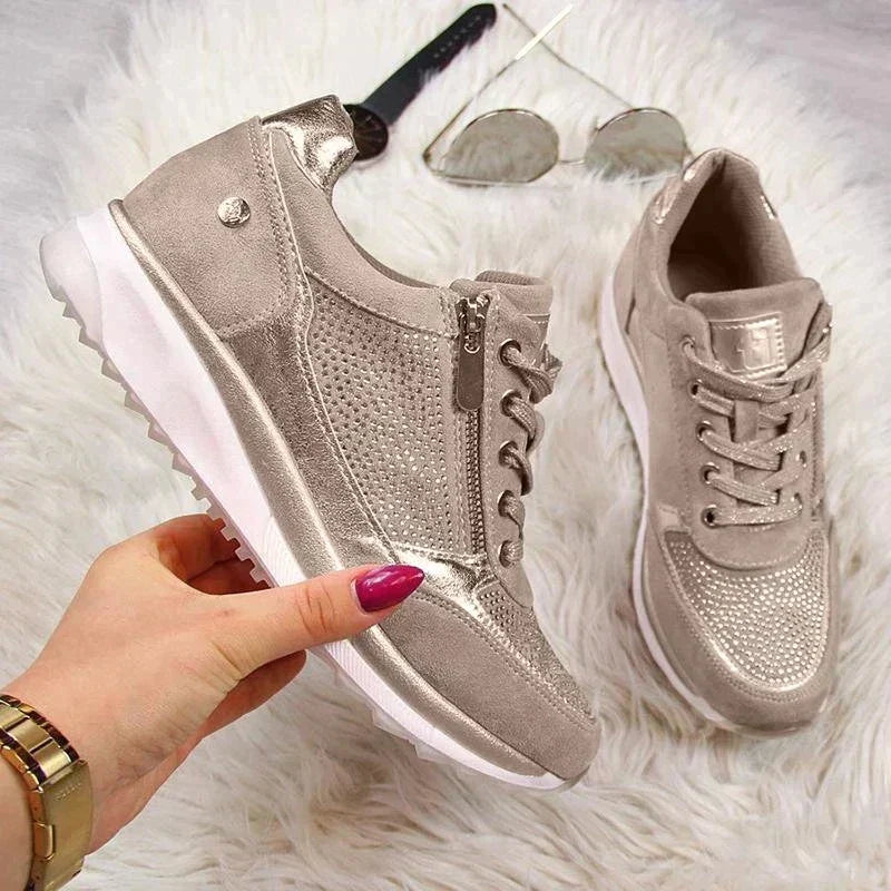 Hand houdt een taupe sneaker met zijrits vast, met de tweede schoen en een zonnebril op wit bont op de achtergrond. 7