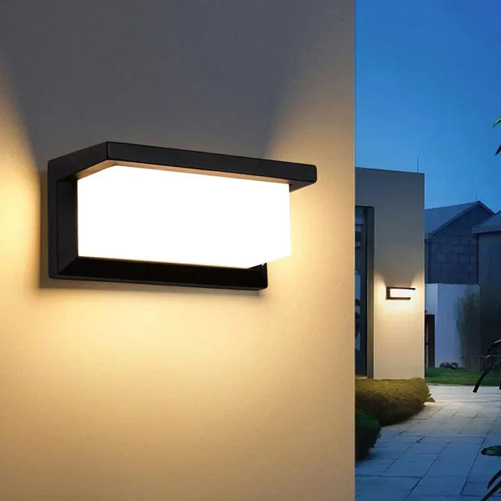 Verlichte wandlamp aan een muur in de schemering, met een tuinpad en huizen op de achtergrond 3