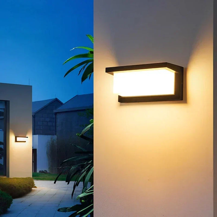 Brandende wandlamp op een lichte buitenmuur, met links een tuinpad, planten en een tweede lamp 4