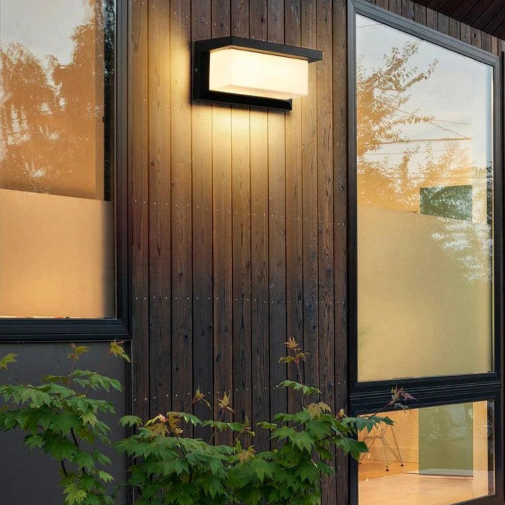 Wandlamp met warm licht tegen een houten gevel tussen twee grote ramen, met struiken eronder 5