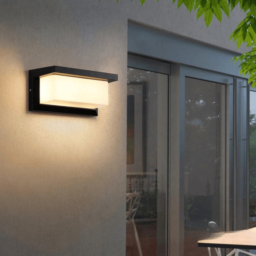 Zwarte rechthoekige wandlamp met warm licht op een grijze buitenmuur naast grote glazen ramen 1