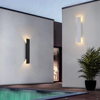 Een zwarte en een witte wandlamp op witte muren naast een zwembad, met een plant ernaast en warm schemerlicht. 4