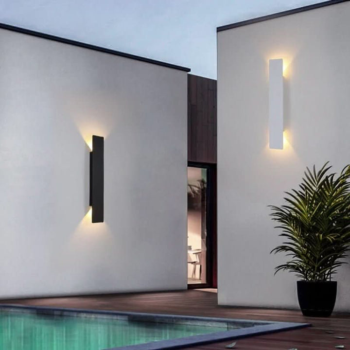 Een zwarte en een witte wandlamp op witte muren naast een zwembad, met een plant ernaast en warm schemerlicht. 4