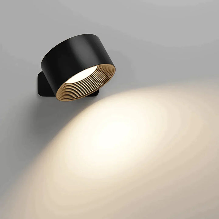 Zwarte wandlamp met ribbels in de binnenkant, schuin gericht en brandend, met een zachte lichtcirkel op de muur. 2
