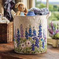 Crèmekleurige gewatteerde mand met geborduurde blauwe en paarse bloemen op een houten vensterbank, gevuld met knuffelhaas en zachte kleding. 1