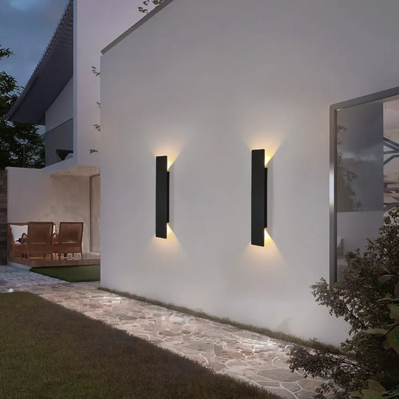 Twee zwarte wandlampen op een witte buitenmuur in de avond, met warme lichtstralen aan boven- en onderkant, naast een terras en tuinpad. 5