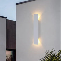 Witte uitvoering van de langwerpige wandlamp op een buitenmuur, met warm licht omhoog en omlaag en een plant in de hoek. 8