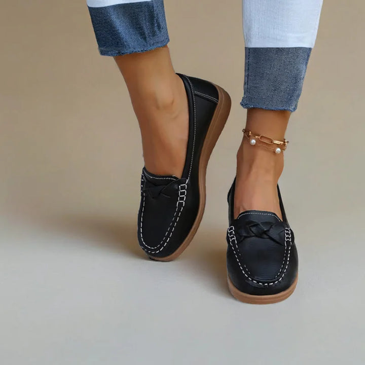 Zwarte loafers met witte stiksels gedragen met opgerolde spijkerbroek en enkelbandje, tegen een beige achtergrond. 3