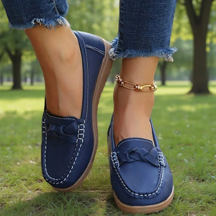 Donkerblauwe loafers met witte stiksels en gevlochten detail, gedragen door een model in een zonnig park. 6