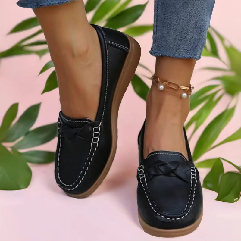 Zwarte instaploafers met witte stiksels en gevlochten detail, gedragen door een model tegen een roze achtergrond met groene bladeren. 2