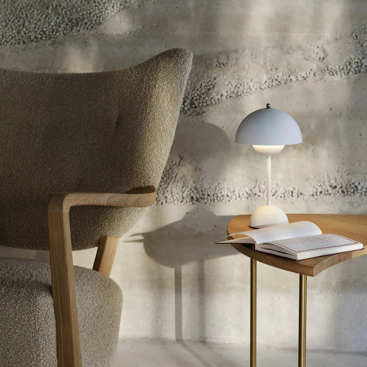 Lichtgrijze mat afgewerkte lamp op een bijzettafel naast een fauteuil, met een opengeslagen boek 16