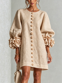 Voorkant van beige mini-jurk met pofmouwen, rij bloemknopen en grote rozenmanchetten, gedragen met pareloorbellen en handtasje. 23