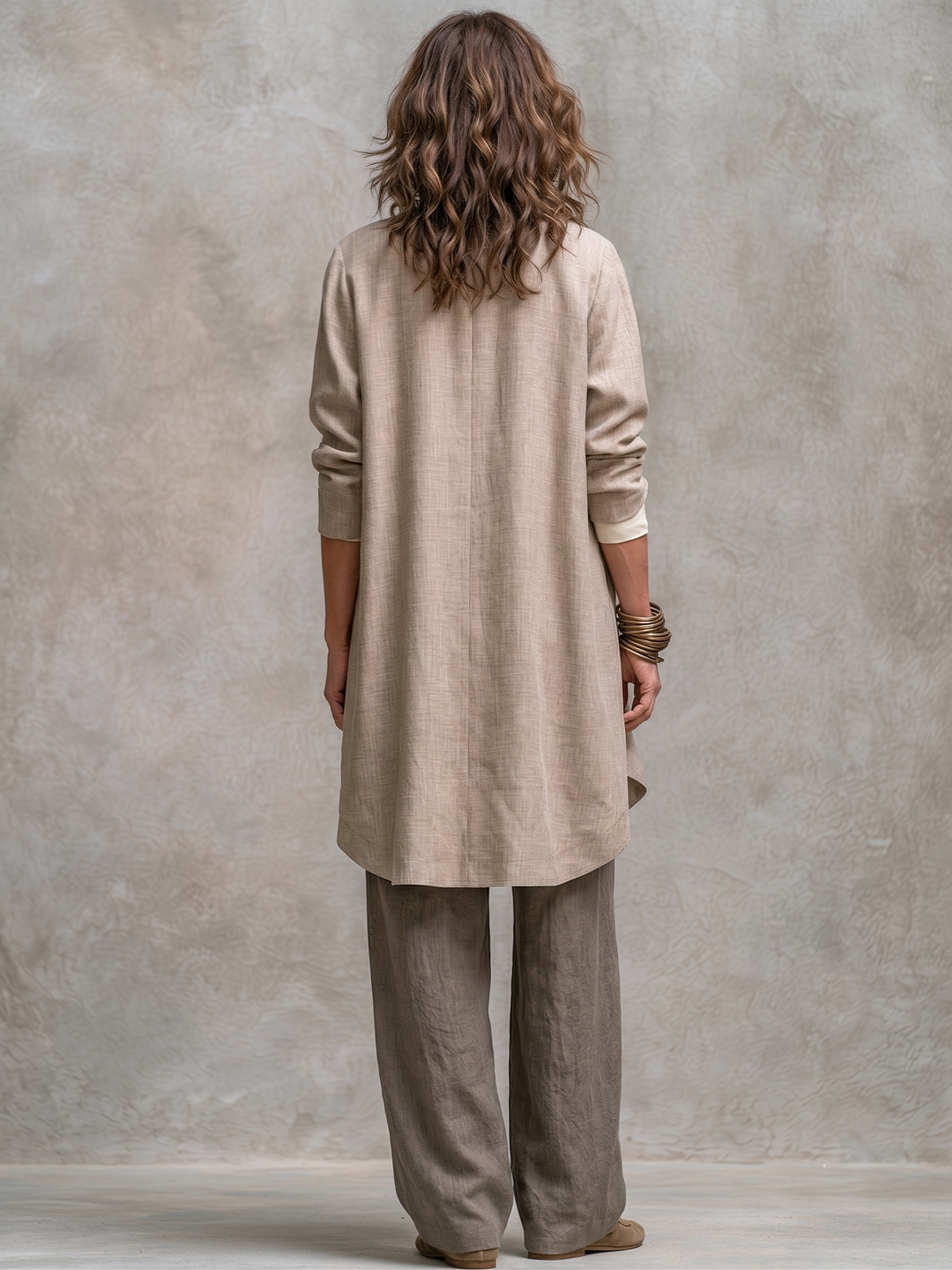Achterkant van de beige lange jack met afgeronde zoom, gedragen over de taupe broek met wijde pijpen. 6