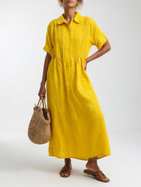 Model in de gele uitvoering, met een hand op de heup, rieten tas in de andere hand en bruine sandalen. 6
