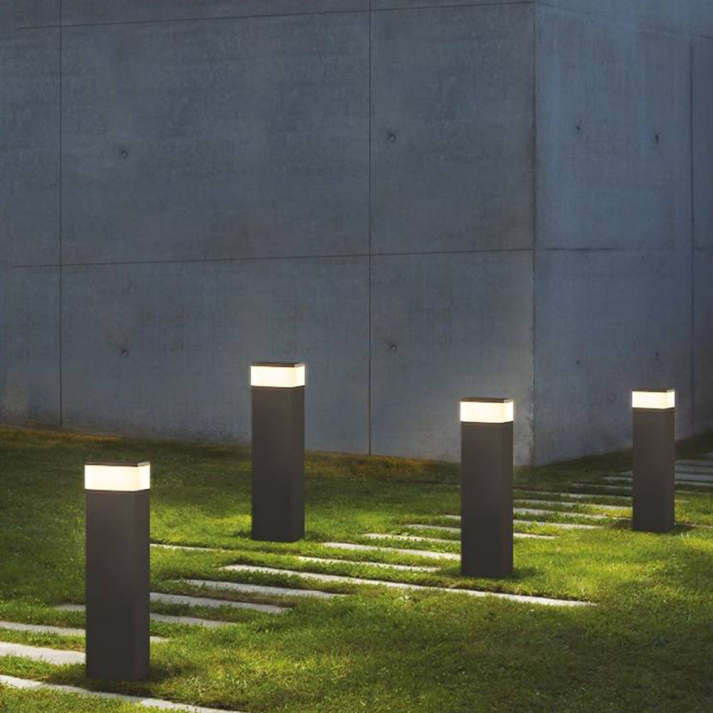 Vier zwarte lichtpalen met warmwit licht langs een pad van tegels in het gras, voor een grijze betonnen muur. 5
