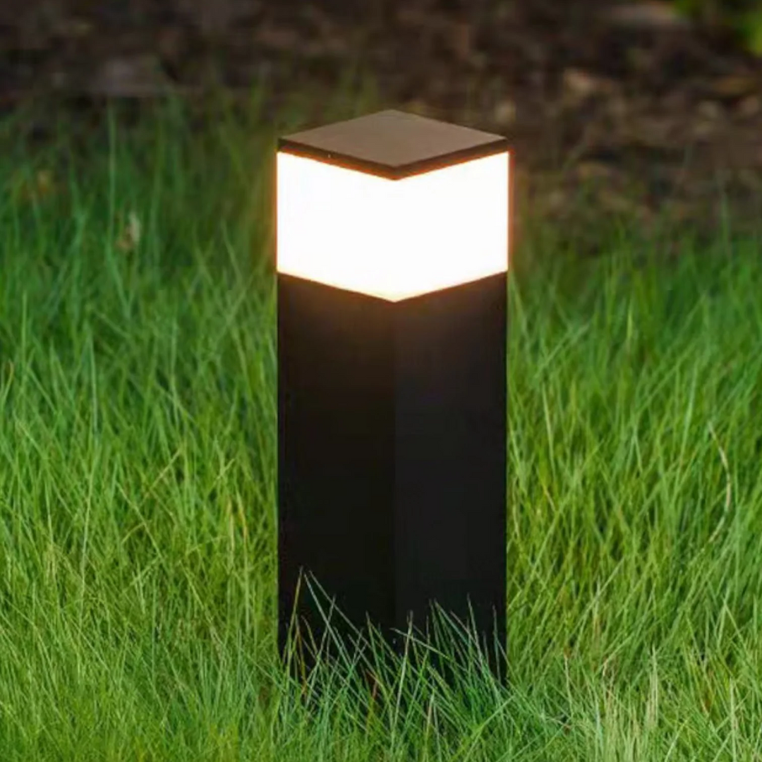 Close-up van een zwarte lichtpaal met helder warmwit oplichtend bovenstuk, staand in hoog groen gras in de schemering. 7