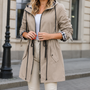 Model in beige jas met trekkoord in de taille, klepzakken en geruite mouwvoering, gedragen met witte broek 3