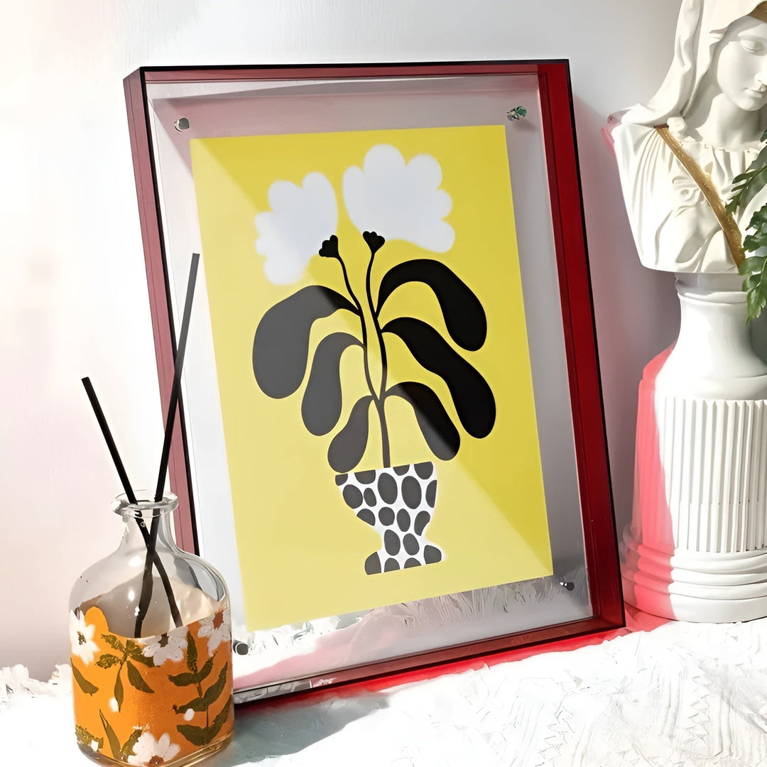 Rode acryl lijst met een gele print van een plant in gestippelde vaas, naast een geurdiffuser en een wit beeld. 15