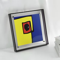 Vierkante acryl lijst met zwarte rand en abstracte print in geel, blauw en rood, tegen een lichte muur. 6