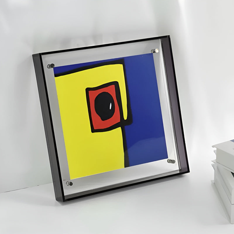 Vierkante acryl lijst met zwarte rand en abstracte print in geel, blauw en rood, tegen een lichte muur. 6