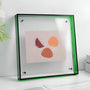 Doorzichtige acryl fotolijst met groene rand en zichtbare bevestigingsschroefjes, staand op een witte tafel met een abstract kunstwerk erin. 21