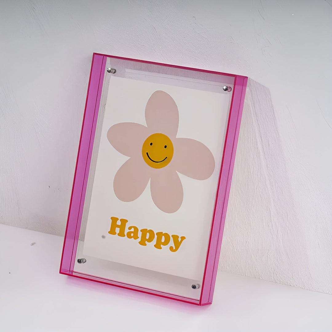 Roze acryl lijst schuin tegen een witte muur, met een bloem met lachend gezichtje in het midden. 19