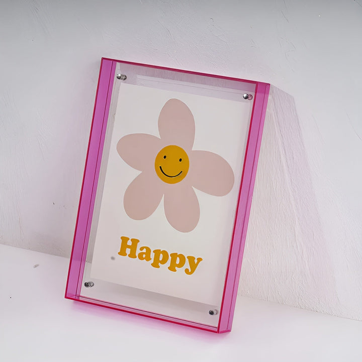 Roze acryl lijst schuin tegen een witte muur, met een bloem met lachend gezichtje in het midden. 19