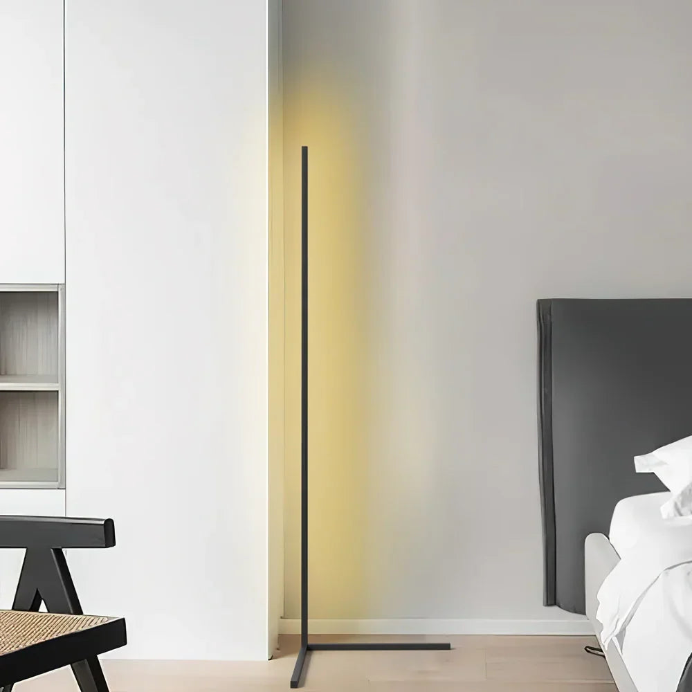 Zwarte vloerlamp met T-voet staat in de hoek van een lichte kamer, met bed en rotan stoel zichtbaar 7