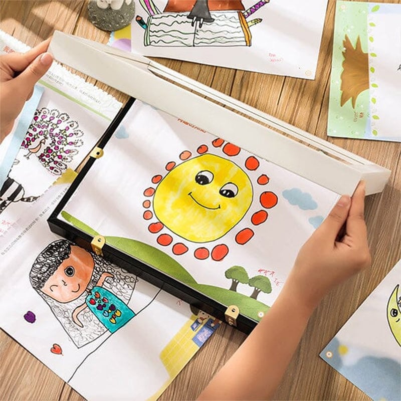 Handen openen een frame boven een tafel vol kindertekeningen, waaronder een lachende zon, om tekeningen te wisselen. 12