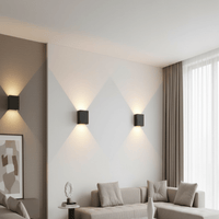 Drie zwarte kubusvormige wandlampen aan een lichte muur in een woonkamer, die licht omhoog en omlaag laten schijnen boven de bank. 1