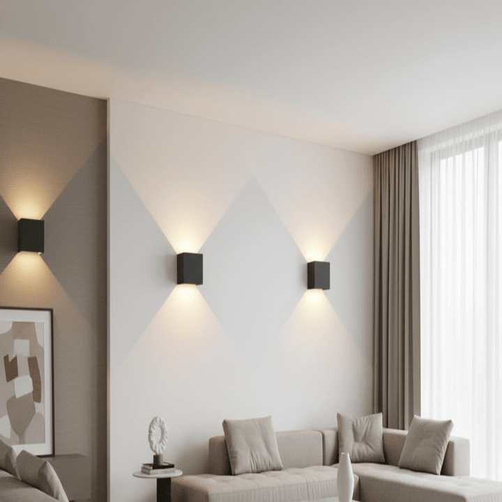 Drie zwarte kubusvormige wandlampen aan een lichte muur in een woonkamer, die licht omhoog en omlaag laten schijnen boven de bank. 1