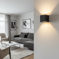 Zwarte kubusvormige wandlamp op de voorgrond met een warme lichtbundel naar boven en beneden, met een woonkamer met grijze hoekbank op de achtergrond. 2