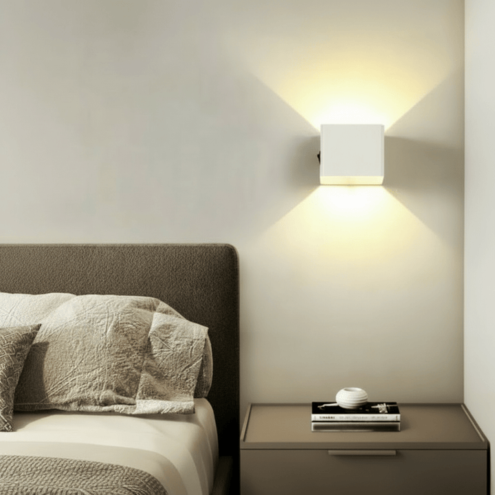 Witte kubusvormige wandlamp naast een bed boven een nachtkastje, met warm licht dat omhoog en omlaag uitstraalt. 3