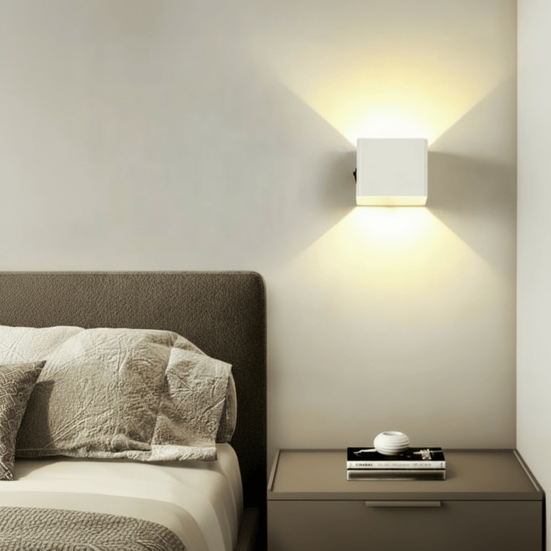 Witte kubusvormige wandlamp naast een bed boven een nachtkastje, met warm licht dat omhoog en omlaag uitstraalt. 3