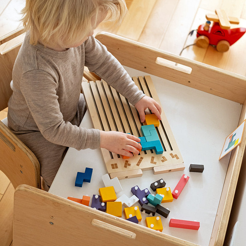 Kind aan een houten kindertafel legt blokken op het puzzelbord, met losse gekleurde stukken en een kaart op het tafelblad. 8
