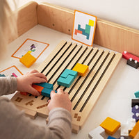 Kind legt met beide handen gekleurde blokken in de groeven van het houten bord, met opdrachtkaarten ernaast. 3